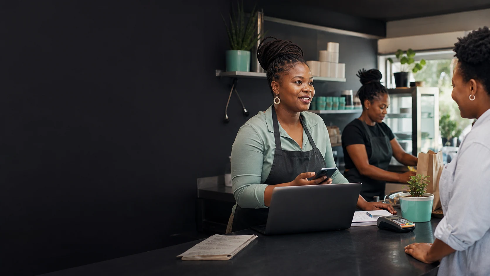 A South African small-business owner helping a customer at a working counter with a laptop and card terminal